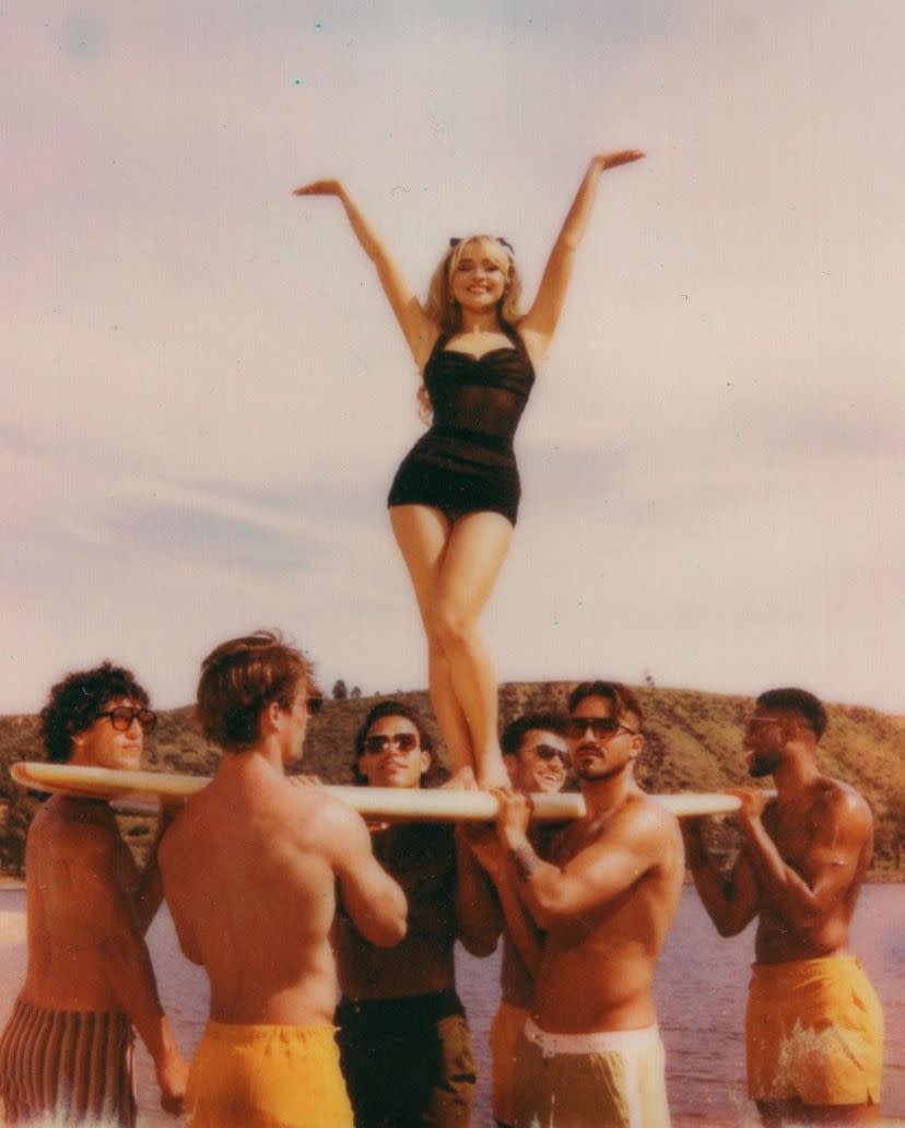 Sabrina Carpenter, in a bathing suit near a body of water, poses on a surfboard held aloft by a half dozen people wearing swimming trunks and sunglasses.