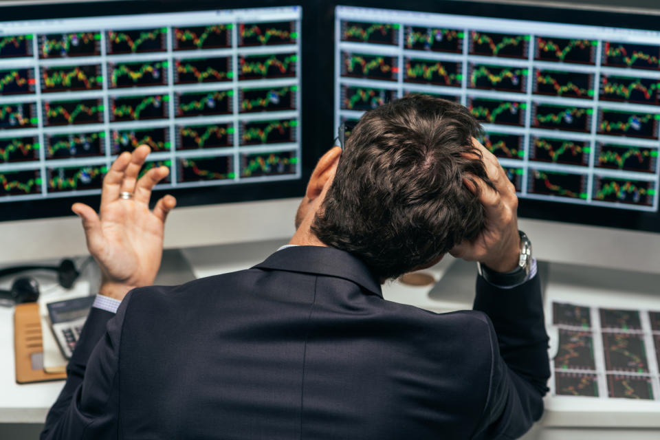 A visibly frustrated businessman in a suit grasping his head as he looks at two computer screens full of stock charts.