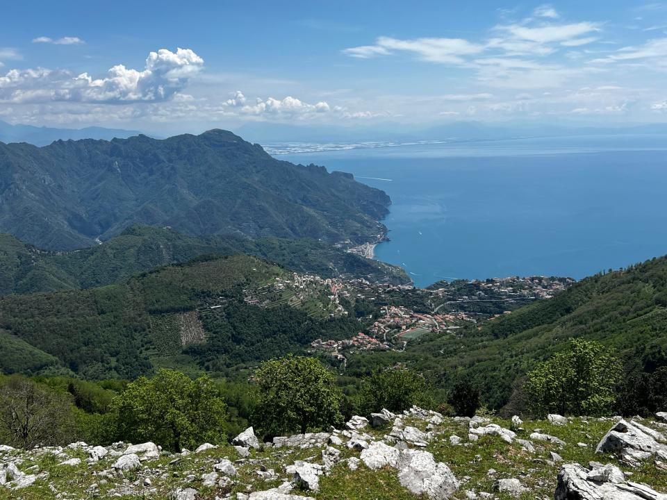 The view from a mountaintop. Greenery-covered mountains and buildings can be seen.