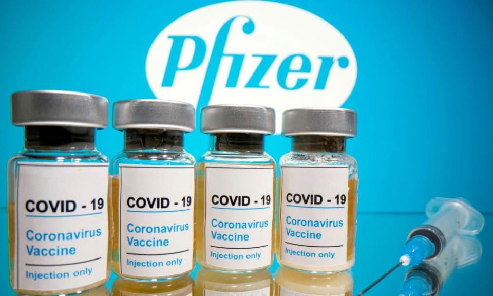 Vials with a sticker reading, “COVID-19 / Coronavirus vaccine / Injection only” and a medical syringe are seen in front of a displayed Pfizer logo.