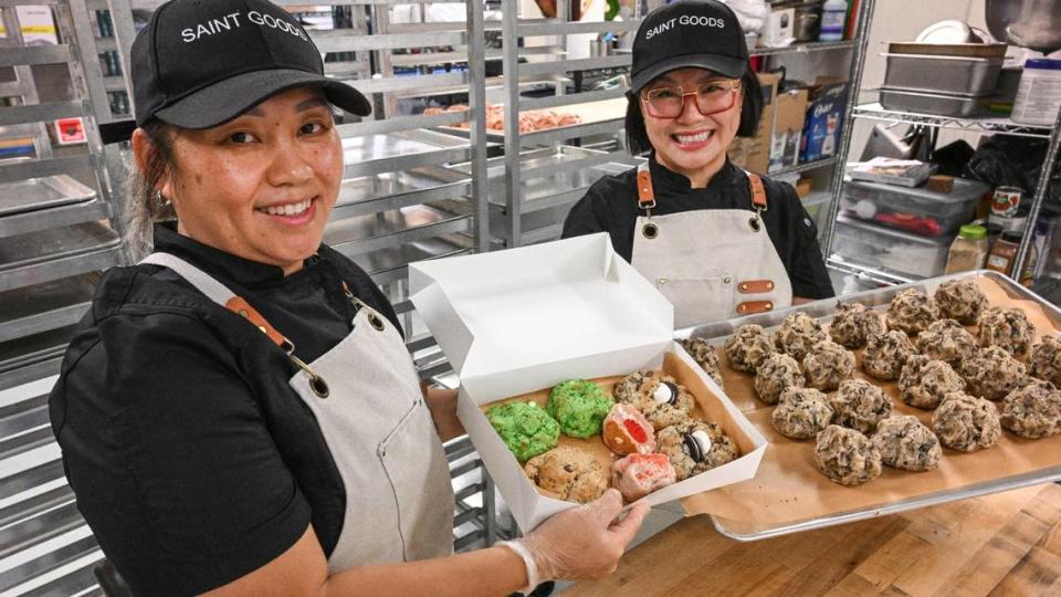Suzy Vang Lo, left, and her sister Nancy Vang, founders of Saint Goods, hold up some of their finished chunky cookies as well as a batch ready for the oven at their workplace inside the Clovis Culinary Center on Wednesday, Nov. 15, 2023.