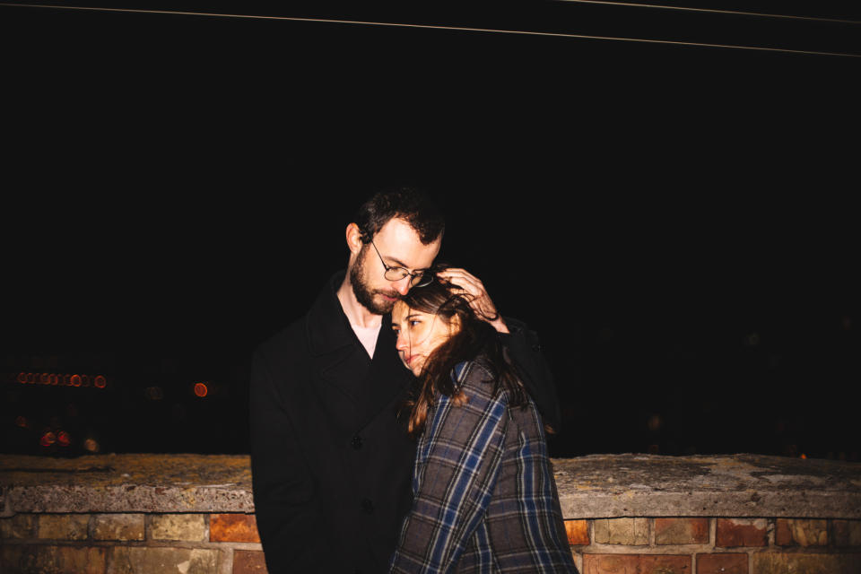 A man and a woman standing close together against a brick wall at night, embracing affectionately, with the woman leaning her head on the man's shoulder