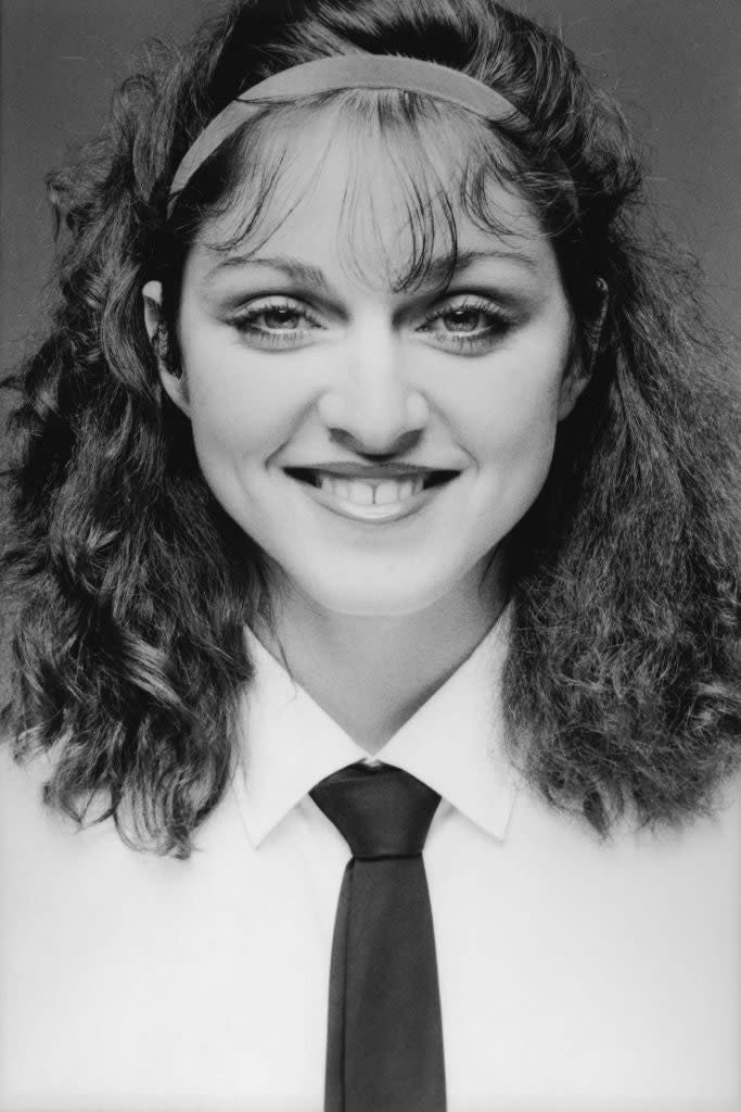 Madonna in the 1980s, smiling at the camera, wearing a white shirt, a black tie, and a headband