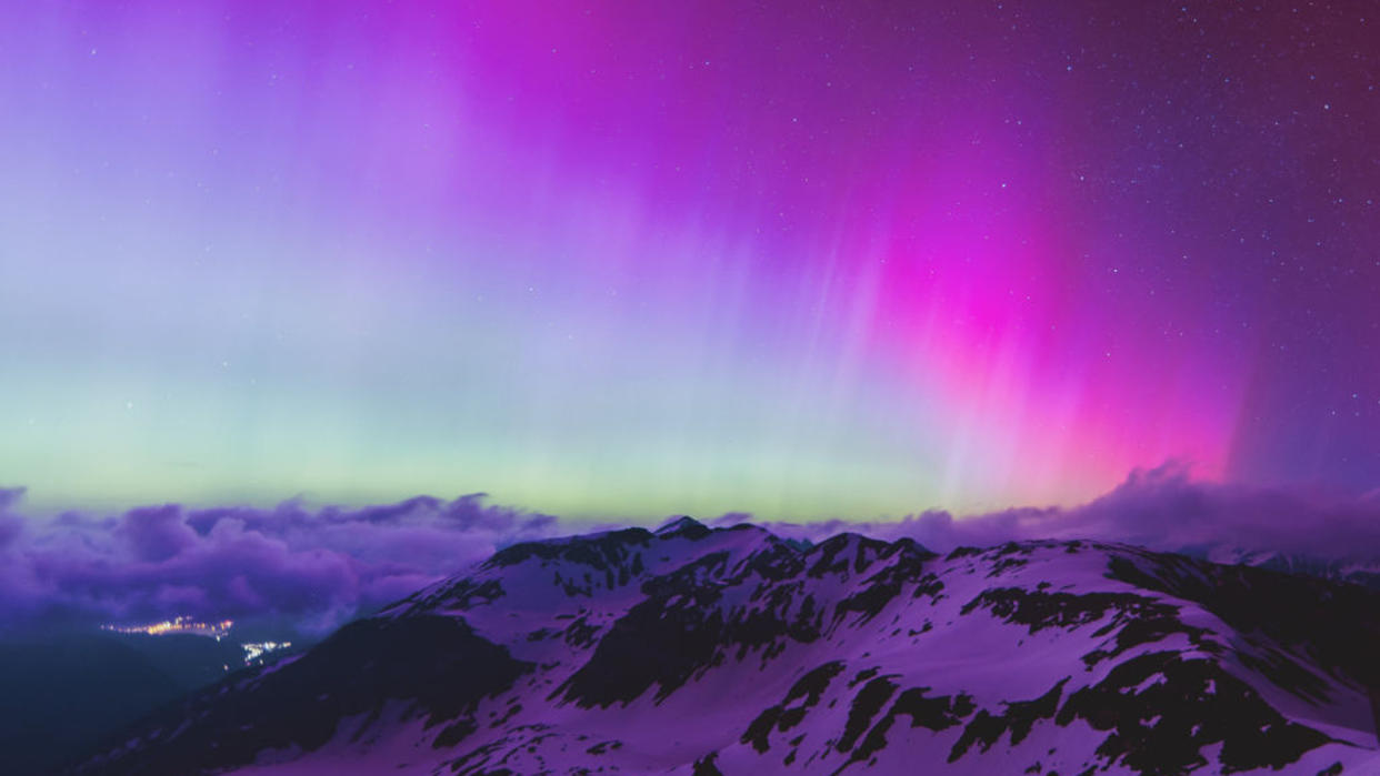  Purple and blue auroras in the night sky above clouds and a mountain. 