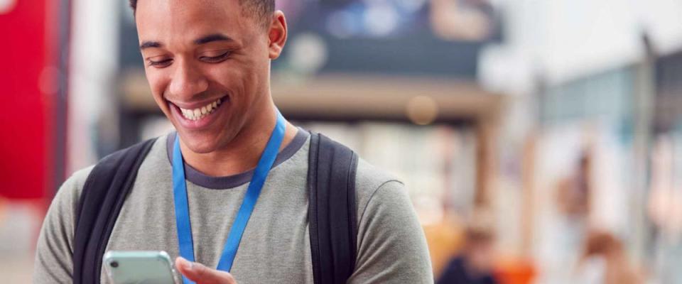 Smiling Male College Student Checking Mobile Phone In Busy Communal Campus Building