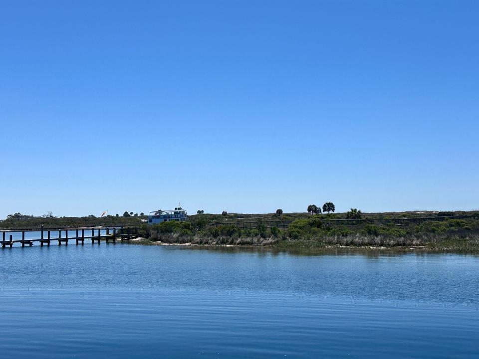 An island full of green grass and trees with a dock and a ferry docked on its beach. Bright-blue water is in the foreground
