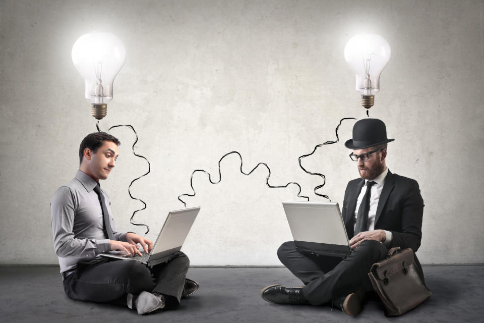 Two people sitting on a floor using their laptops. Lit-up lightbulbs shine above their heads.