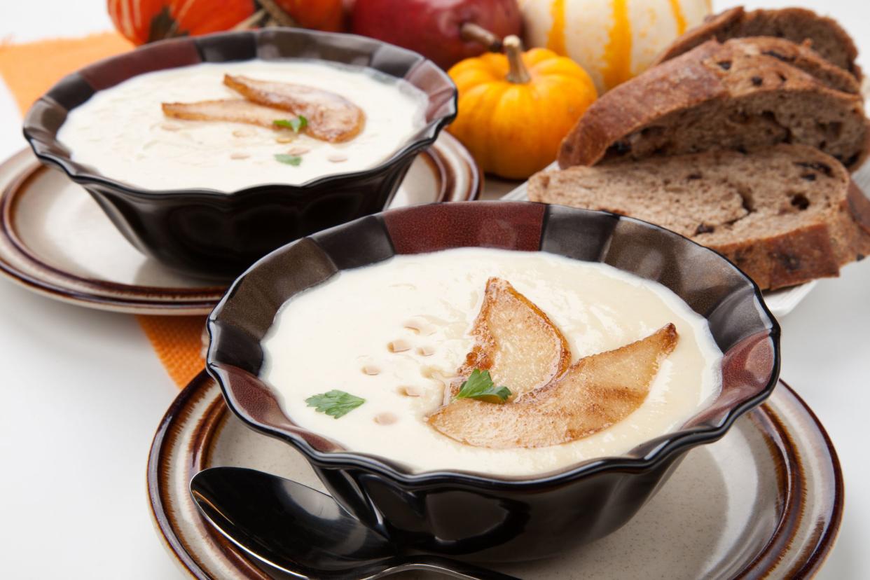 Two bowls of hot delicious Roasted Parsnip and Pear Soup. Garnished with slices of caramelized pears, maple syrup, and parsley. Pumpkins and bread..