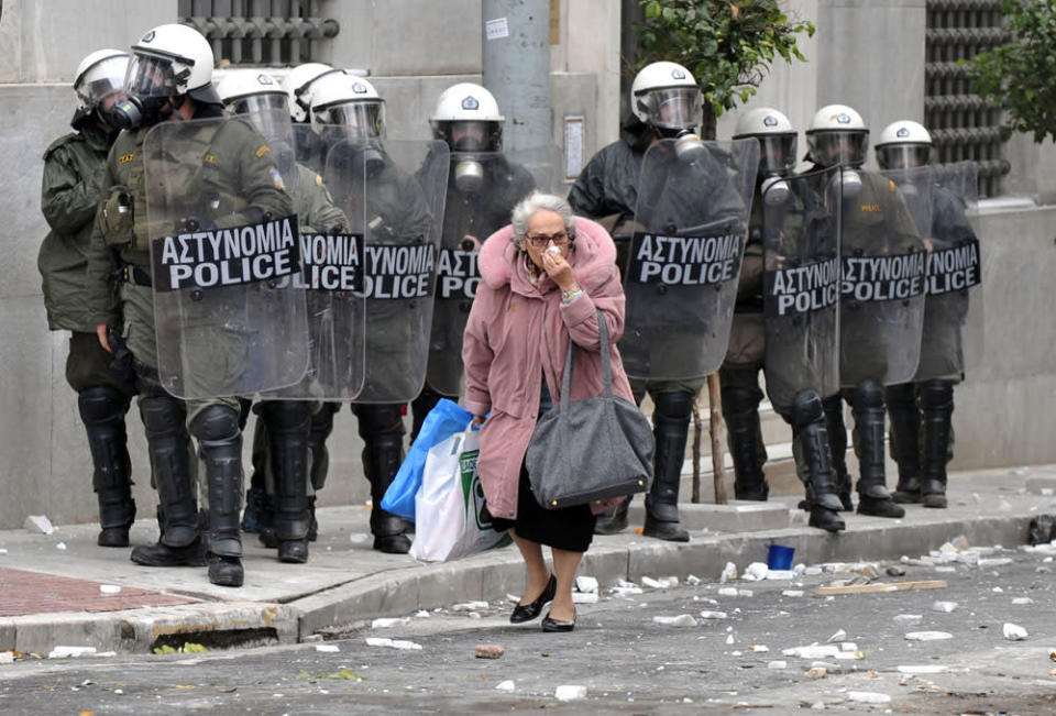 Le pensioni sono state tagliate di oltre il 20% e con la poca assistenza gli anziani sono una delle categorie che ha subito le maggiori ripercussioni. Finendo nel calderone di quel 40% della popolazione che ha varcato la cosiddetta “soglia di povertà”. <br> Nella foto un'anziana costretta a girare per le strade di Atene coprendosi il volto per proteggersi dal gas lacrimogeno lanciato poco prima dalle forze dell'ordine in tenuta anti-sommossa.