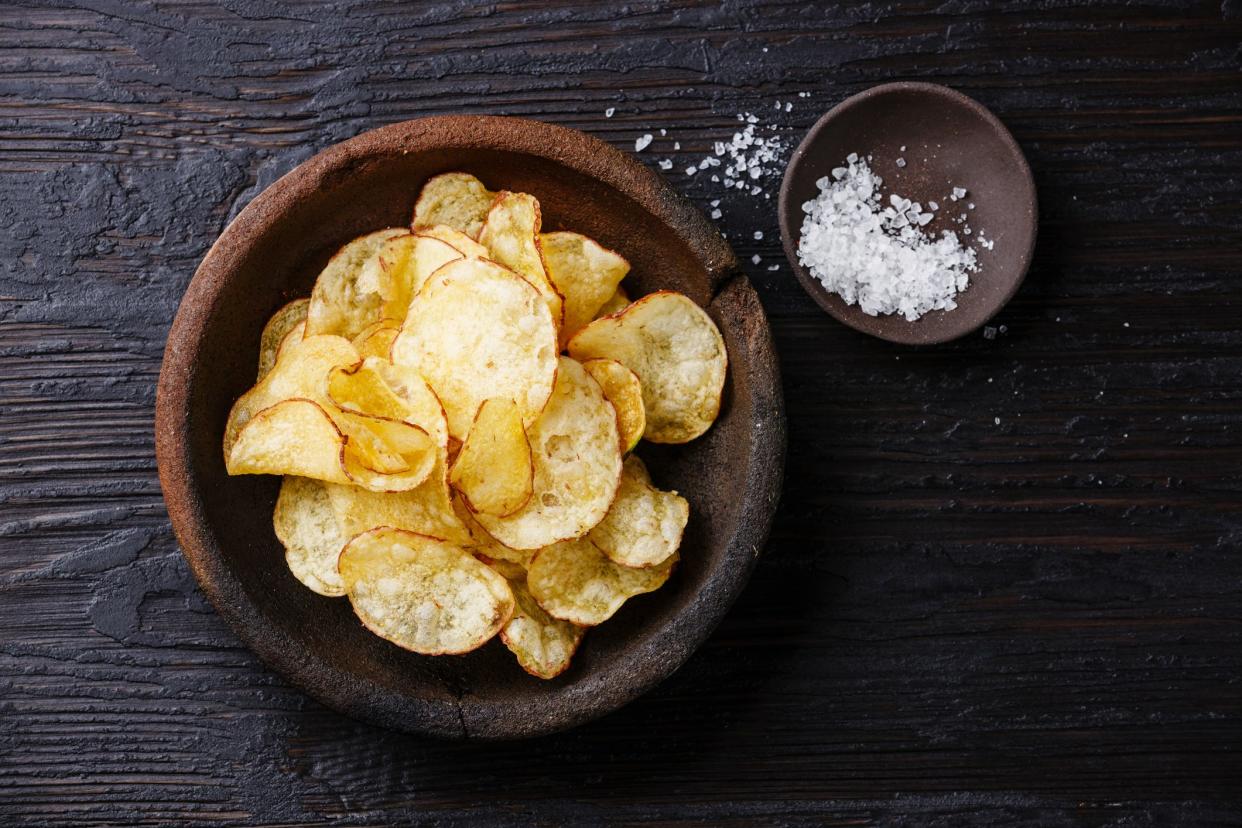 Potato chips in a wooden bowl near a small wooden bowl of powdered white salt on a dark wooden table with gradient shadowing