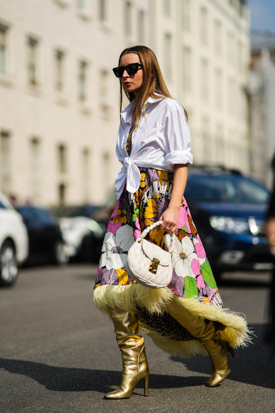 MILAN, ITALY - SEPTEMBER 23: Julia Comil wears black sunglasses, gold earrings, a gold chain necklace, a white knotted shirt with rolled-up sleeves, a yellow / green / brown / white / pink / black / flower print pattern shoulder-off / asymmetric long dress with pale yellow feathers on the borders, a white shiny ruffled leather handbag from Miu Miu, gold shiny ruffled leather pointed heels knees boots, gold rings, outside the Etro fashion show during the Milan Fashion Week - Spring / Summer 2022 on September 23, 2021 in Milan, Italy. (Photo by Edward Berthelot/Getty Images)