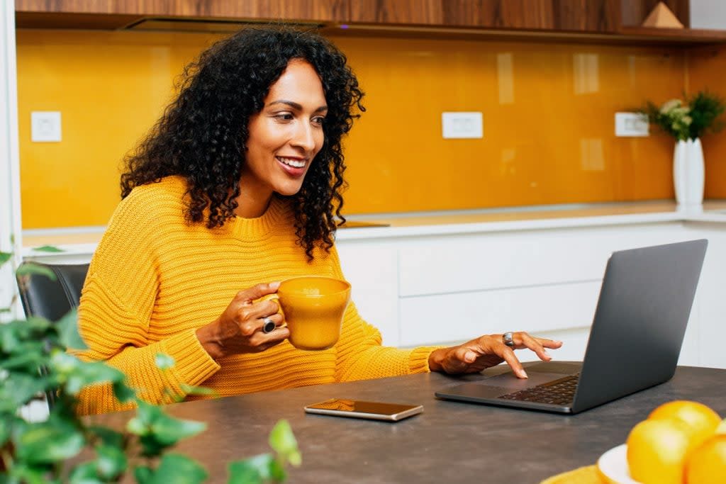 A woman smiles as she scrolls on the internet using her laptop. She's wearing a yellow sweater and holding a yellow coffee mug. 