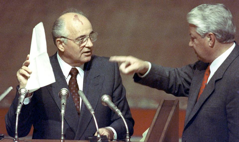 A man wearing a suit and tie and glasses holds up papers before microphones as another man, right, points to them