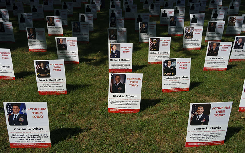 A photo display of held up military appointments is seen outside the Capitol. A large number of signs are planted in the grass, extending into the distance. On the signs are the names and photos of military members whose appointments are being held up. Each sign also reads "#CONFIRMTHEMTODAY" in the upper right corner.