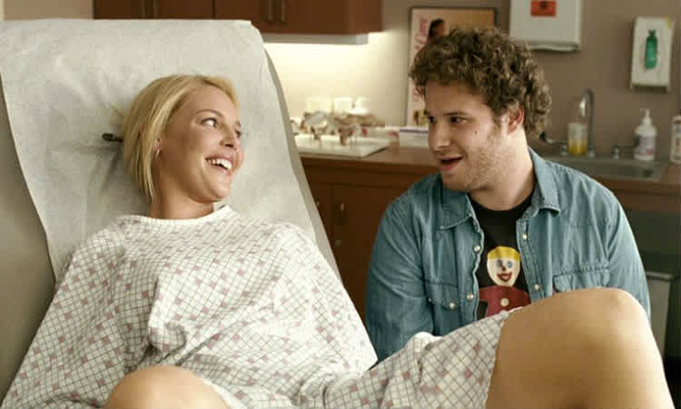 A woman on an exam table smiles at a man standing beside her.