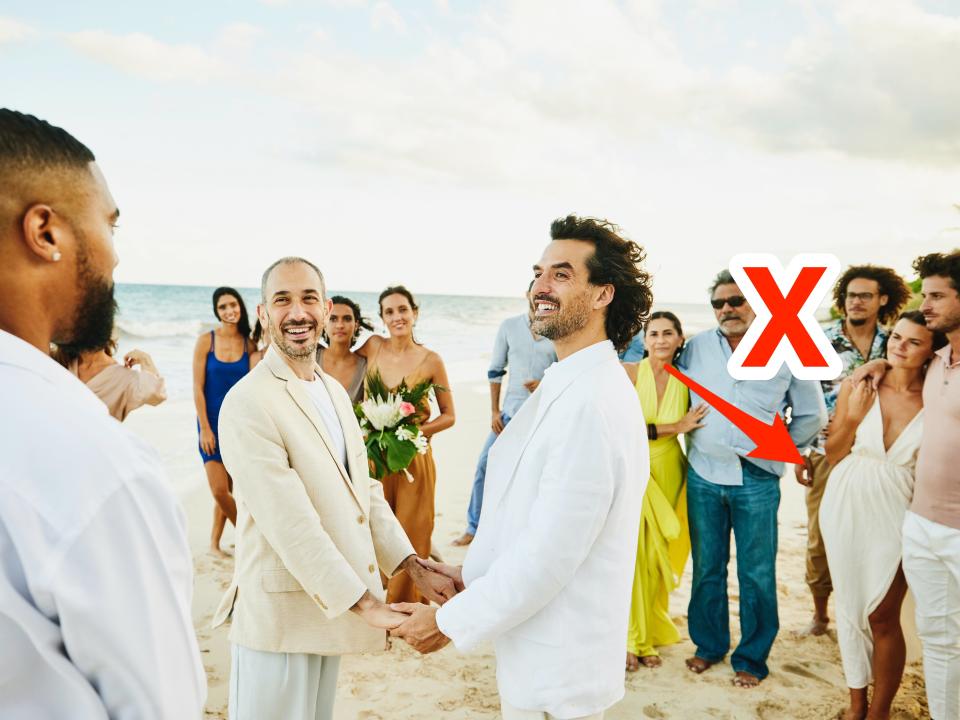 A couple holds hands on the beach during their wedding. An arrow and an x point at a woman wearing white.