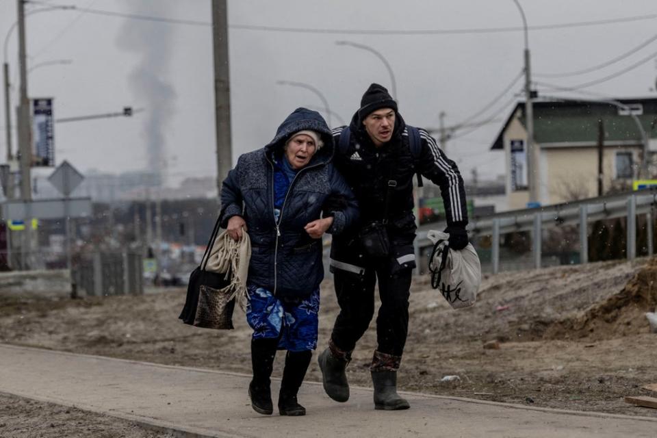 A man helps an elderly woman to run for cover after heavy shelling on the only escape route used by locals, while Russian troops advance towards the capital, in Irpin, near Kyiv, 6 March 2022 (REUTERS)