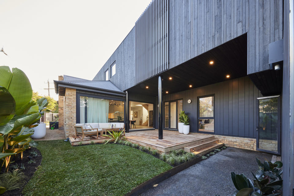 A wide shot of the front of the house with black cladding and a wooden deck. A grass on the right. 