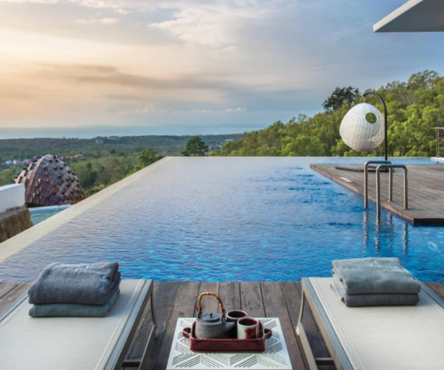 Looking straight over a vibrant blue infinity pool that looks out to the green landscape of Ulutwatu with the ocean in the far distance. 