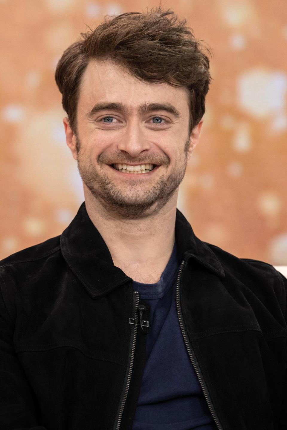 Daniel Radcliffe smiling, wearing a dark jacket and blue shirt, seated on a couch