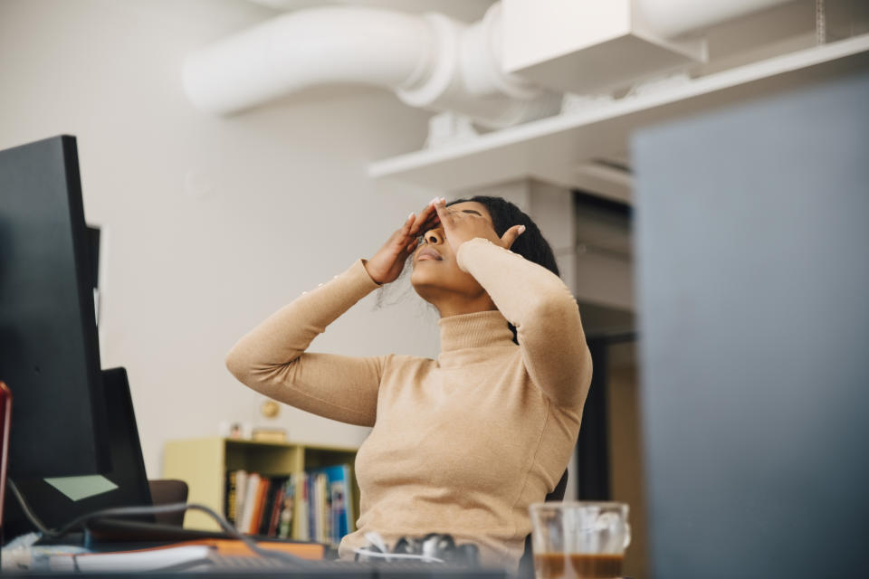 Person in a beige turtleneck sits at a desk in an office space, leaning back with hands over their eyes, appearing stressed or tired