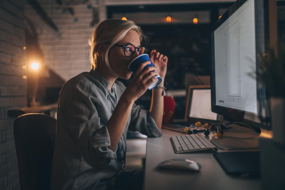 A person with glasses works late at night, sipping from a cup while looking at a computer screen. The room is dimly lit with a cozy atmosphere