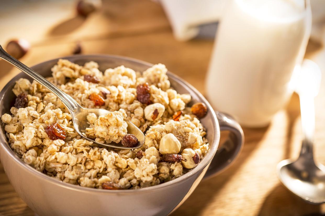 Closeup of nutty granola with a spoon in a beige mug, on the left side, on a wooden table, with a blurred background of a glass of milk, a spoon, and more items