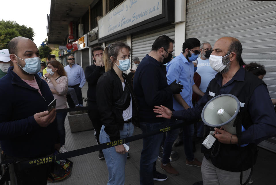 A money transfer employee, right, holds a megaphone as he asks citizens who wear masks and gloves to help curb the spread of the coronavirus, outside a Western Union shop to receive their money transfer in U.S. dollar currency, during the last day that they are allowed to dispense dollars to customers following new Central Bank rules, in Beirut, Lebanon, Thursday, April 23, 2020. (AP Photo/Hussein Malla)