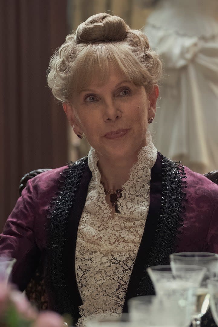 Christine Baranski, portraying Agnes van Rhijn, in a Victorian outfit with lace details, sits at a dining table in a period-piece setting