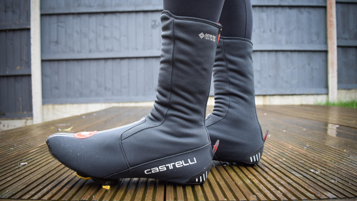  A close up of a black pair of Castelli Estremo overshoes worn by a cyclist who is stood on a wooden deck 