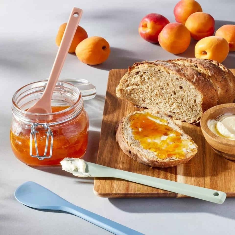 Jar of jam with pink spatula, mint green spatula, blue spatula, sliced bread with jam, and fruit on a table