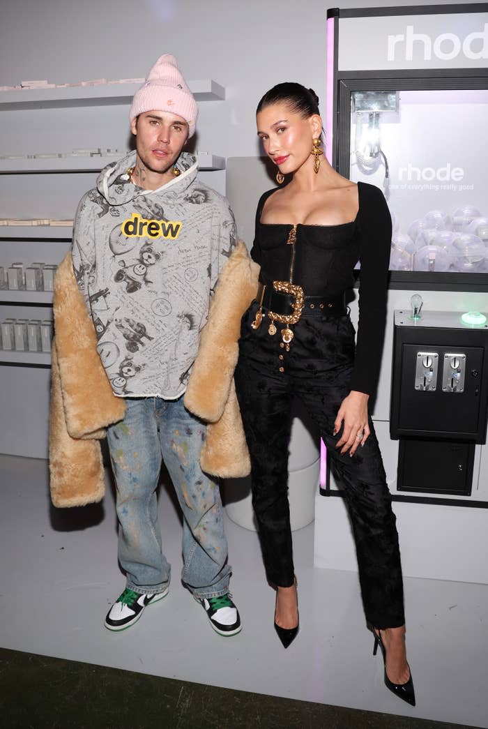 justin dressed in paint-splattered jeans, oversized hoodie, and beanie while hailey looks polished in an all-black outfit, heels, and gold accessories