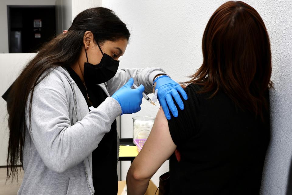 A medical worker, wearing a black face mask and blue rubber gloves, administers an injection into the upper arm of someone whose back is to the camera.
