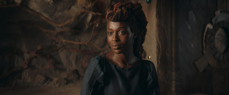 A woman with long braided hair stands in a dimly lit, rustic setting, wearing a simple dark dress