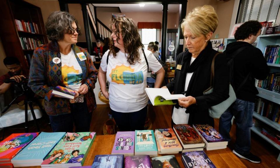 Three white woman talk and stand before a table holding piles of books.