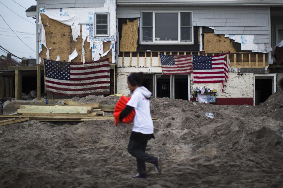 A volunteer passes a damaged home emblazoned with U.S. flags in the Rockaways, Saturday, Nov. 10, 2012, in the Queens borough of New York. Despite power returning to many neighborhoods in the metropolitan area after Superstorm Sandy crashed into the Eastern Seaboard, many residents of the Rockaways continue to live without power and heat due to damage caused by Sandy. (AP Photo/John Minchillo)