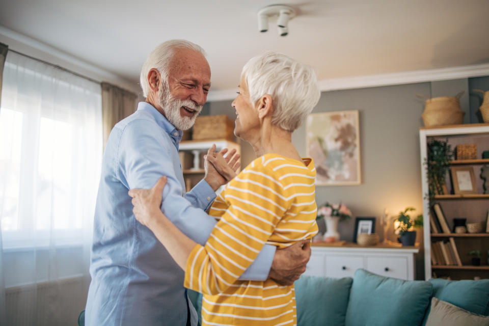 An elderly couple is smiling and dancing in their living room, holding each other closely and gazing into each other's eyes