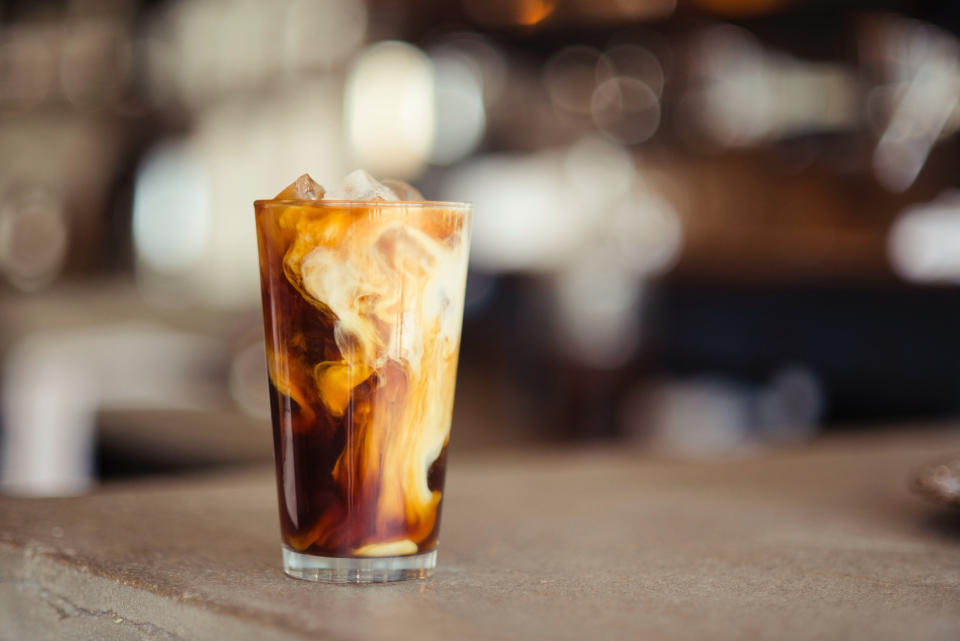 A glass of iced coffee with cream being poured in, creating swirls, on a counter