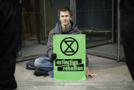 An Extinction Rebellion climate change protester with his hand glued to the floor outside the London Stock Exchange holds a banner in the City of London, Thursday, April 25, 2019. The non-violent protest group, Extinction Rebellion, is seeking negotiations with the government on its demand to make slowing climate change a top priority. (AP Photo/Matt Dunham)