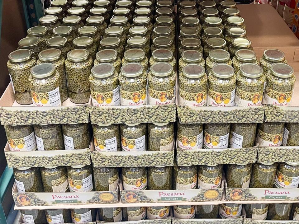 Large jars of capers with labels depicting a woman. Jars are placed in cardboard boxes with metal lids
