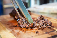 Shredded meat on a cutting board with tongs pulling it apart