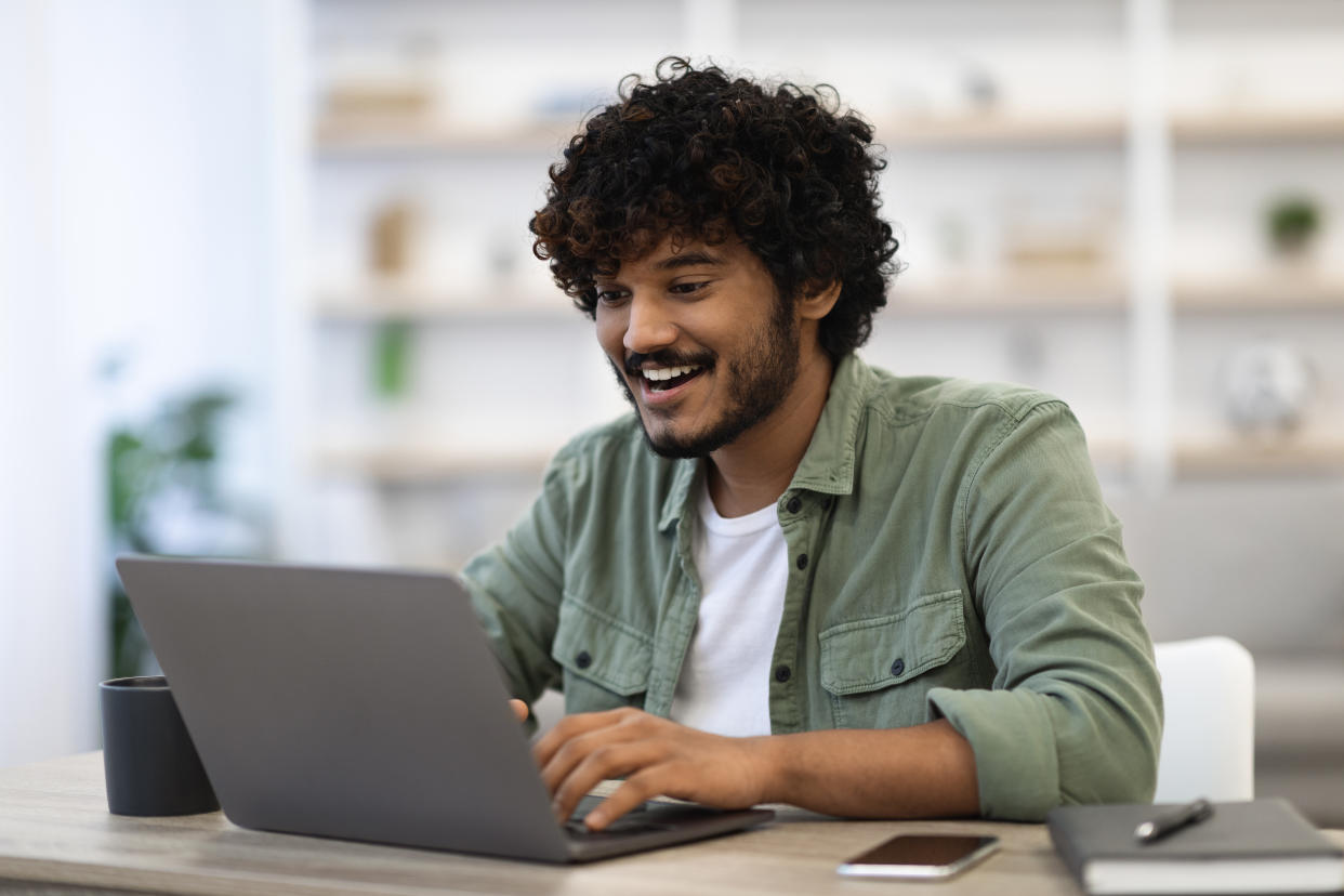 Happy dark-skinned curly bearded young man in casual sitting at workdesk at home, using modern laptop, typing on computer keyboard and smiling, looking for job online, copy space