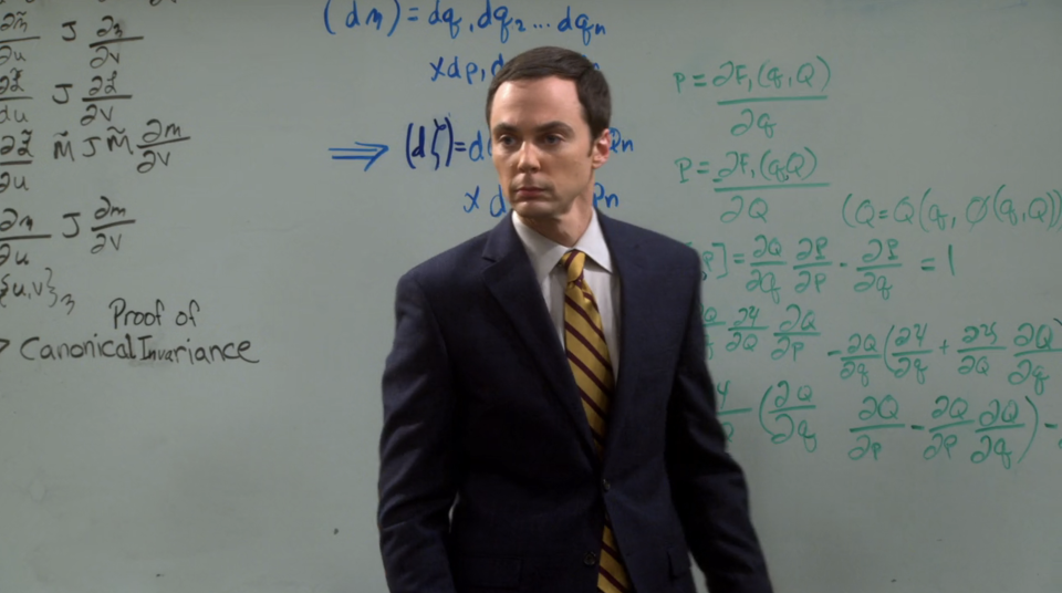 Sheldon Cooper, in a suit and striped tie, stands in front of a whiteboard filled with complex mathematical equations