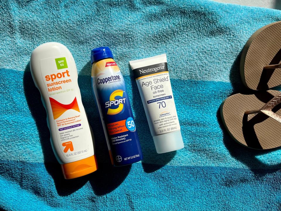 Three bottles of various sunblock on a beach towel with flip flops off to the side.