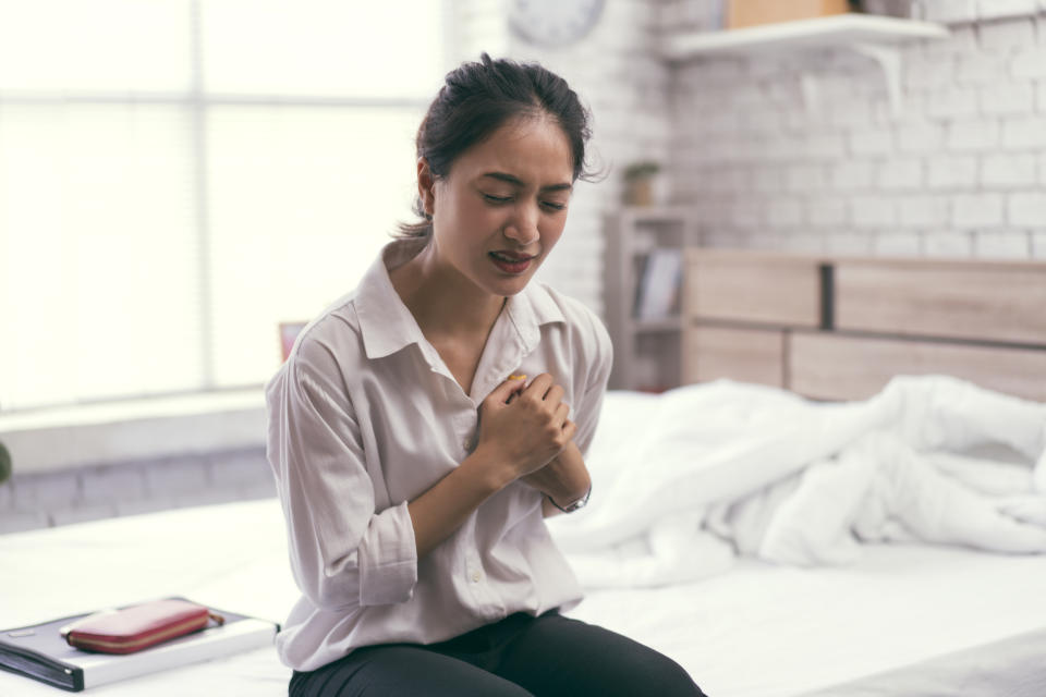 Women wears a white shirt and black pants. She sits on a bed and is clutching her chest with a pained expression on her face.