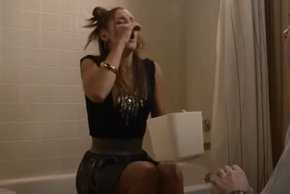 A woman sits on the edge of a bathtub, appearing to cry or wipe her eyes, holding a tissue box. A standing man is partially visible