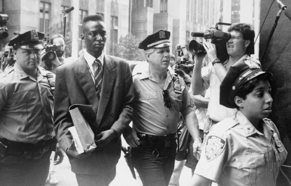 Yusef Salaam, in suit and tie, is flanked by NYPD officers, as a news reporter points a video camera at him.