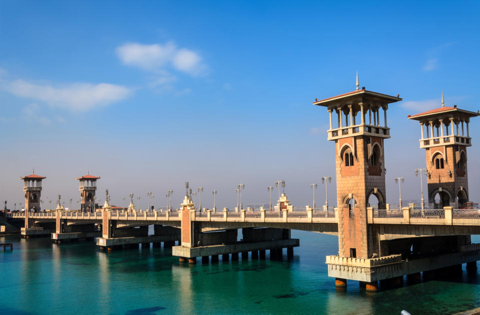 The image shows the Montaza Bridge with its distinctive towers and architectural design spanning over clear blue water under a partly cloudy sky