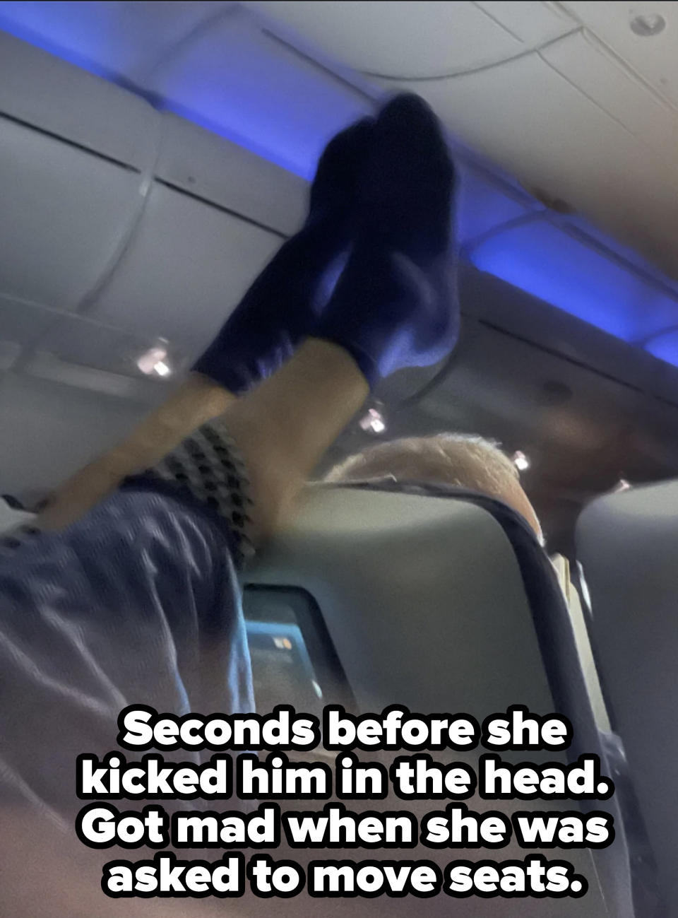 A person's legs are resting on an airplane seat, with feet in socks extending above the seatback. The cabin ceiling and lights are visible in the background