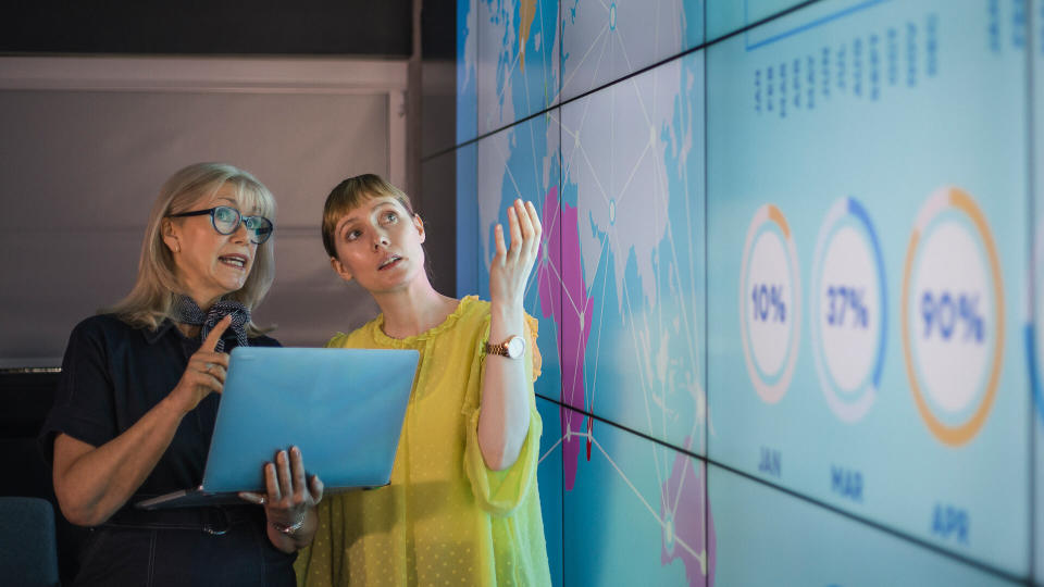 An experienced woman mentors a female colleague, the mature woman is holding a laptop as they debate data from an interactive display; they are both wearing smart casual clothing.